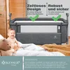 A baby sitting next to a crib with a teddy bear. Produktbild-Vorschau