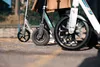 Close-up of three scooters parked on a paved surface, with people's legs visible behind them. Produktbild-Vorschau