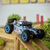 A remote-controlled off-road truck is positioned on a wooden ramp, with a blurred background of greenery and red posts. Produktbild-Vorschau
