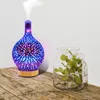 An aroma diffuser with a starburst pattern and colored lights is shown on a wooden surface next to a glass vase with green leaves. Voir les images du produit