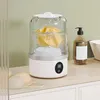A mini washing machine with a yellow garment inside, placed on a countertop next to a soap dispenser. Produktbild-Vorschau