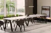 A modern dining room with a long white table and chairs, set in front of large windows overlooking a garden. Produktbild-Vorschau