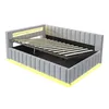 A light gray upholstered bed frame with the mattress support mechanism raised, revealing storage space underneath. Yellow LED lighting is visible along the bottom and headboard. Produktbild-Vorschau