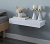 A white floating shelf mounted on a light gray wall, next to a bed. The shelf holds decorative items. Náhled obrázku produktu