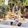 A group of four people are enjoying an outdoor gathering, toasting with drinks around a white folding table laden with food and decorations. Náhľad obrázku produktu
