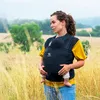 A woman is holding a baby in a sling in a field of tall grass. Produktbild-Vorschau