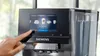 A hand is shown interacting with the touchscreen display of a Siemens coffee machine, selecting a beverage option. Produktbild-Vorschau
