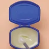 An open jar of Vaseline with a spatula inside, against a light peach background. Podgląd zdjęcia produktu