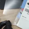 A person wearing black gloves is holding a clear protective film over a wooden surface, with packaging visible to the right. Produktbild-Vorschau