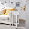 A white side table with a drawer and shelf sits next to a sofa with yellow and cream pillows. A glass of drink is on top of the table. Produktbild-Vorschau