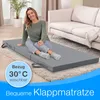 A woman is sitting on a folded mattress in a living room. Produktbild-Vorschau