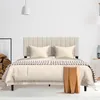A bed with a beige headboard and bedding is shown in a room with a wooden stool and a stack of logs. Produktbild-Vorschau