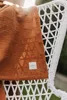 A close-up of a textured brown blanket with a geometric pattern, draped over a white wicker chair outdoors. Produktbild-Vorschau