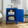 A blue nightstand with its door open, displaying books and a folded blanket inside. A lamp and picture frame are on top. Podgląd zdjęcia produktu