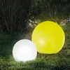 Two illuminated garden spheres, one white and one yellow, sit on a grassy lawn surrounded by tall reeds and bushes. Produktbild-Vorschau