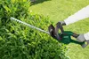A person is using a green hedge trimmer to trim a dense green bush in a garden setting. Produktbild-Vorschau