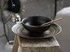 A black bowl with gold trim and a gold spoon rests on a decorative plate, which is placed on a rustic wooden stool. A blurred branch with small white flowers is visible in the background. Náhľad obrázku produktu