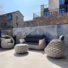 A patio set with a sofa, two chairs, and a coffee table is arranged on a tiled patio in front of a decorative wall. Produktbild-Vorschau