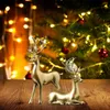 Two golden reindeer figurines are placed on a wooden surface in front of a decorated Christmas tree with blurred lights. Produktbild-Vorschau