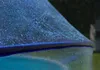 A close-up of a blue, possibly glass or plastic, structure with a textured surface and small bubbles or imperfections. The background is blurred, showing greenery and a darker area that could be foliage or a fence. Produktbild-Vorschau
