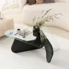 A modern living room scene featuring a unique coffee table with a glass top and a sculptural black base, adorned with a vase of dried flowers, a candle, and a cup of coffee. Produktbild-Vorschau
