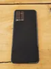 The back of a black smartphone with a textured pattern is shown lying on a wooden surface. Produktbild-Vorschau