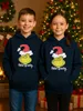 Two children wearing matching Grinch hoodies stand in front of a Christmas tree and fireplace. Produktbild-Vorschau