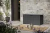 A grey storage box sits on a patio next to a stone wall, with a dog lying on a rug in front of it. Produktbild-Vorschau