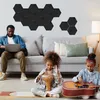 A family is in a living room, with the father working on a laptop on the couch and two children playing music on the floor. Black hexagonal acoustic panels are mounted on the wall. Produktbild-Vorschau