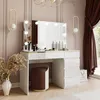 A modern white vanity table with a lighted mirror, makeup, and a stool in a room with pink walls and brown curtains. Produktbild-Vorschau
