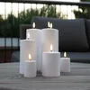 Several white LED candles of varying sizes are arranged on a wooden table outdoors, with a blurred background of outdoor furniture and greenery. Produktbild-Vorschau
