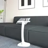A white C-shaped side table with a cup on it is placed next to a dark gray sofa in a living room. Podgląd zdjęcia produktu