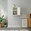 A white wall-mounted cabinet with its door open, displaying a deer figurine and some books. A potted plant is on the floor to the left, and a framed picture is on the floor to the right. Produktbild-Vorschau