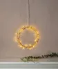 A circular LED light wreath hangs from a thin wire against a textured wall. A vine with green leaves and small white flowers rests on a white shelf below. Podgląd zdjęcia produktu