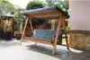 A wooden swing chair with a gray cushion and canopy is set up on an outdoor patio with plants and a building in the background. Produktbild-Vorschau