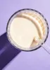 A top-down view of a glass of vanilla protein shake with a straw, set against a light purple background. Produktbild-Vorschau