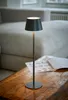 A dark gray LED table lamp with a conical shade sits on a wooden table next to some papers. A sofa is visible in the background. Produktbild-Vorschau