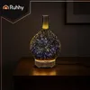 A glass aroma diffuser with a starburst pattern, lit with colorful LED lights, sits on a wooden table in a dimly lit room. Produktbild-Vorschau