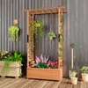 A wooden planter box with a trellis structure, filled with various plants and flowers, set on a wooden deck against a gray wooden wall. Podgląd zdjęcia produktu