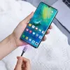 A hand is holding a smartphone while a USB-C cable is being plugged into it. The phone screen displays a colorful wallpaper and app icons. The background is a textured white surface, possibly a blanket or bedspread. Produktbild-Vorschau