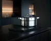 A stainless steel pot with a lid sits on a dark countertop in a dimly lit kitchen. Produktbild-Vorschau