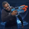 A young person wearing safety glasses and a hoodie holds a blue and orange Nerf Elite 2.0 Commander RD-6 blaster, aiming it forward. The background features a futuristic, digital grid pattern with glowing blue circles. Anteprima dell’immagine prodotto