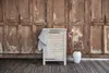 A wooden laundry basket with a towel draped over it, placed on a wooden deck in front of a rustic wooden door. A vase with pink flowers is next to the basket. Produktbild-Vorschau