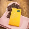 A yellow chocolate bar wrapper and loose chocolate pieces are arranged on a pink cloth and a wooden surface. Produktbild-Vorschau