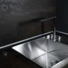 A close-up view of a modern kitchen sink and faucet with a dark, textured backsplash. Produktbild-Vorschau