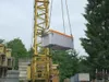 A yellow crane is lifting a large gray and orange toolbox outdoors. Produktbild-Vorschau