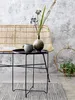 A stylishly arranged vignette featuring a small black side table with a grey mug and a decorative vase with dried flowers, set against a textured wall and a woven bench. Produktbild-Vorschau