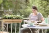 A woman is sitting on a balcony, reading a book, with a planter box on the railing and a table with drinks and plates. Produktbild-Vorschau