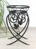 A black metal plant stand with decorative heart-shaped scrollwork and leaf accents, placed on a light-colored floor with a green plant to the left. Náhled obrázku produktu