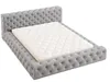 A tufted gray bed frame with a white mattress on top, shown from a diagonal angle against a white background. Náhľad obrázku produktu